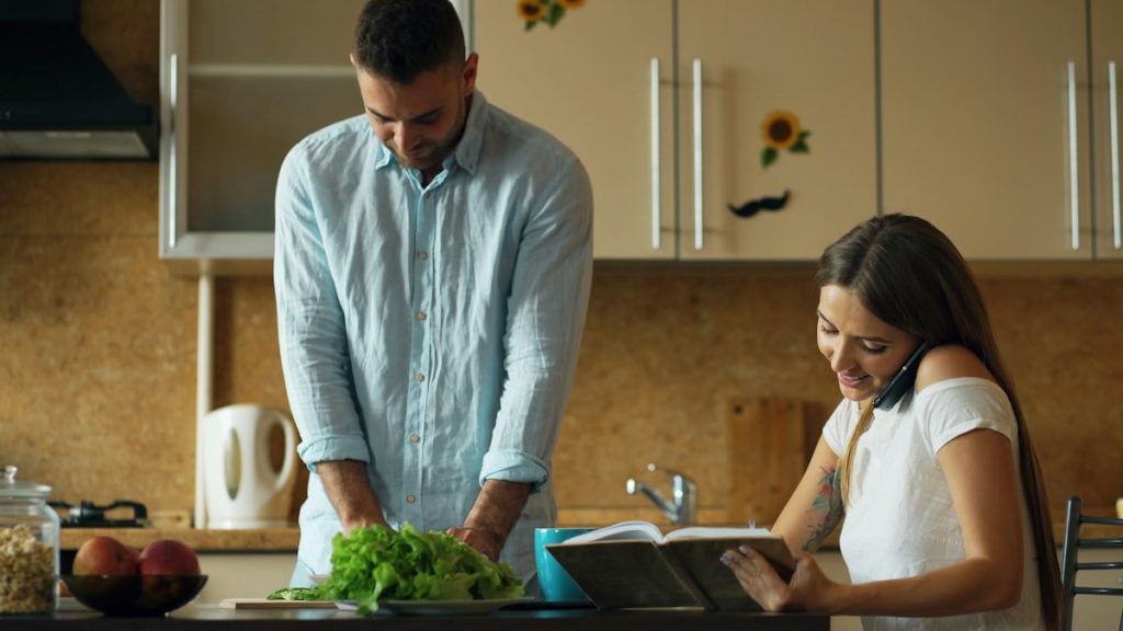 Attractive couple in the kitchen early morning. Beautiful girl chatting phone while her boyfriend cooking breakfast at home